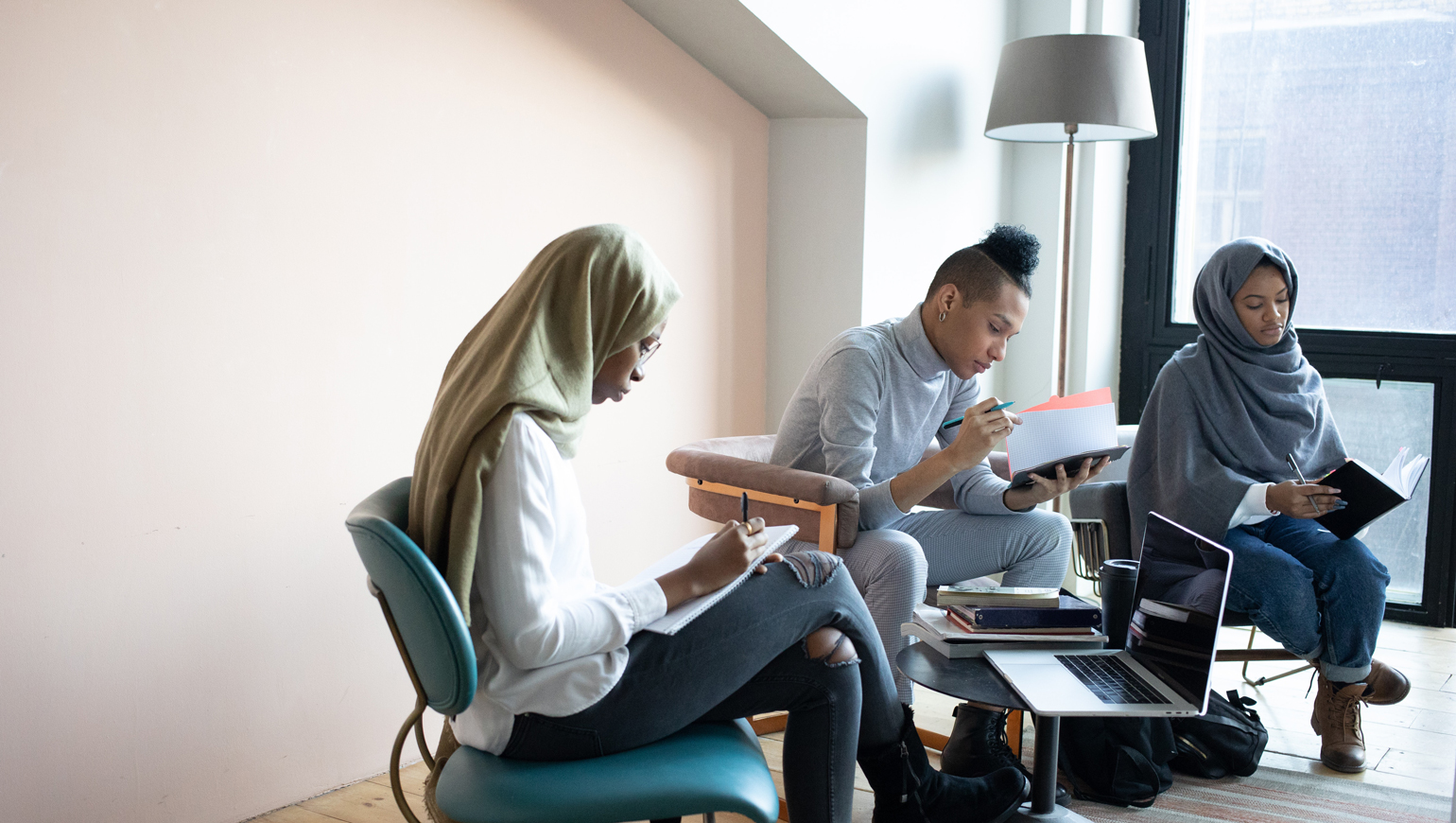 Drie personen zitten op een stoel te lezen en schrijven in een schrift en er staat een tafeltje met boeken en een laptop