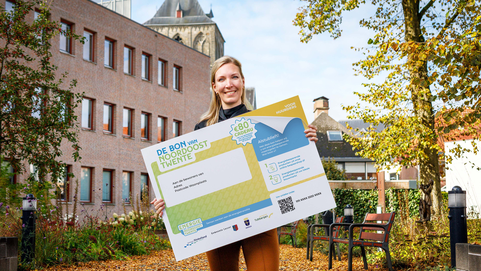 Vrouw houdt grote versie van de bon van Noordoost Twente vast met op de achtergrond een gebouw, een kerk en huizen