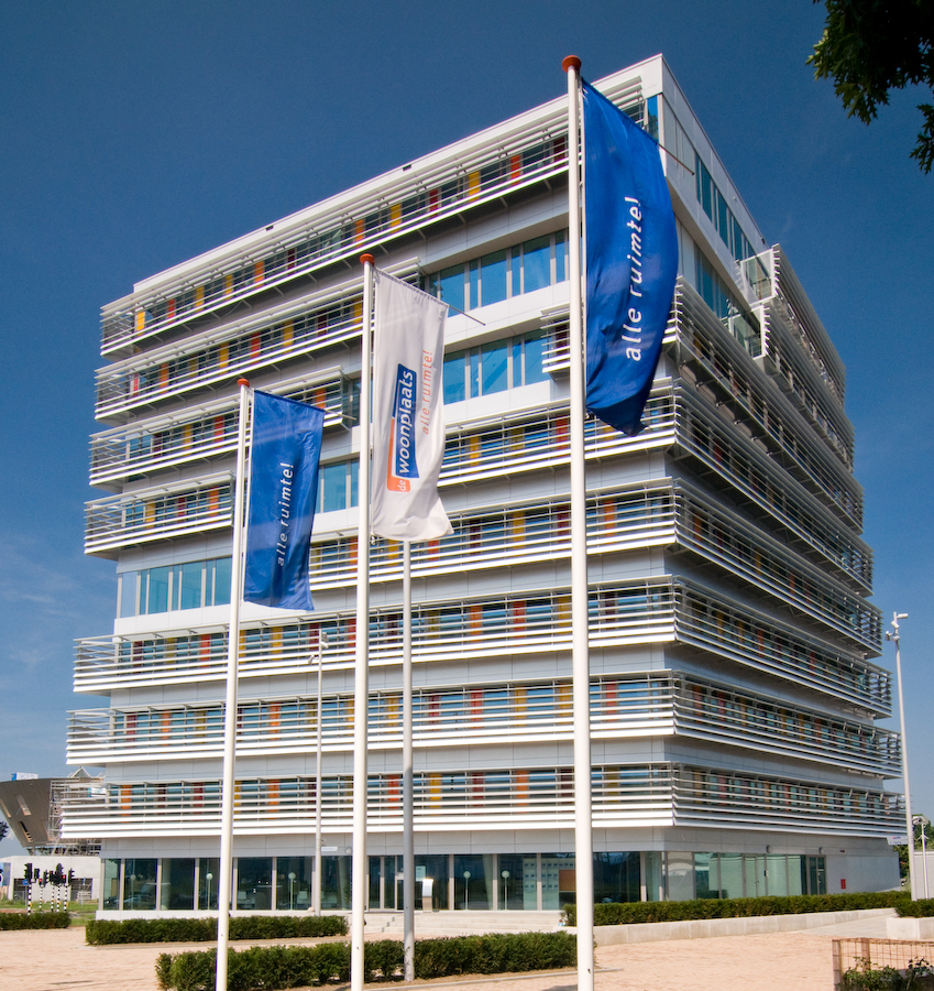 Hoog wit gebouw met daarvoor drie vlaggen, één witte met het logo van De Woonplaats en twee blauwe met de tekst: alle ruimte!