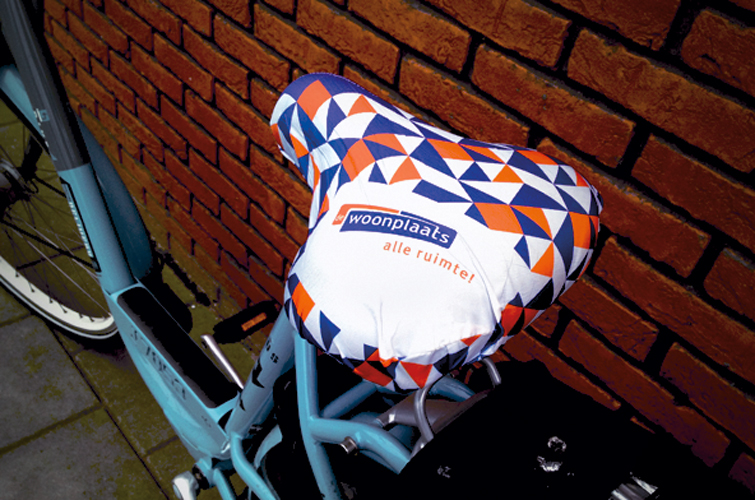 Blauwe fiets met een zadelhoes die de kleuren en het logo van De Woonplaats bevat