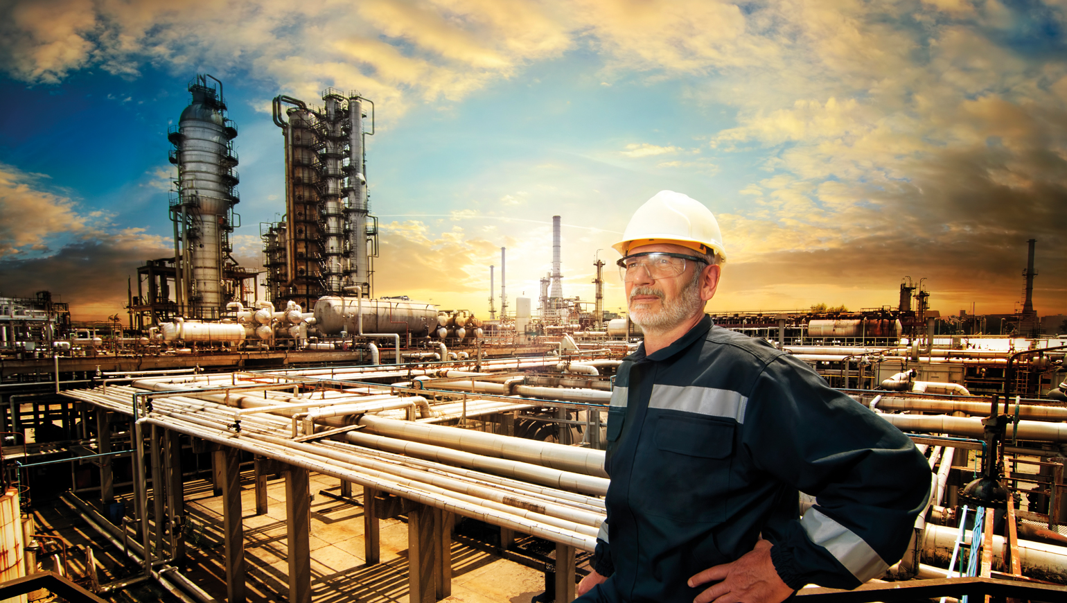 Man in werkpak met bescherm bril en helm op staat voor een fabrieksterrein met buizen en installaties