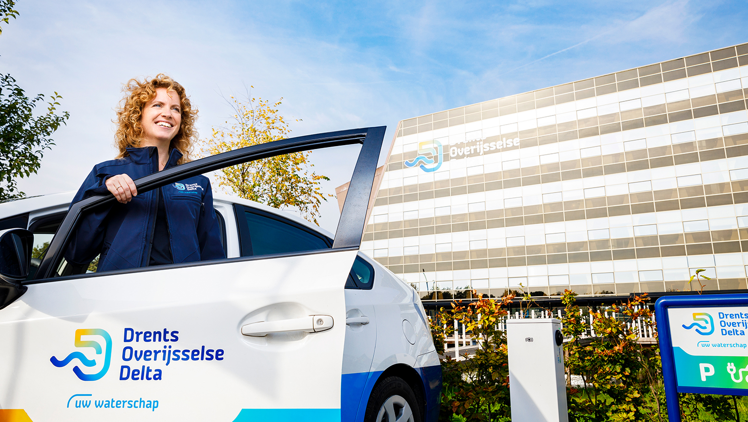 Vrouw stapt uit een witte auto, met Drents Overijsselse Delta logo erop, voor het gebouw van Drents Overijsselse Delta